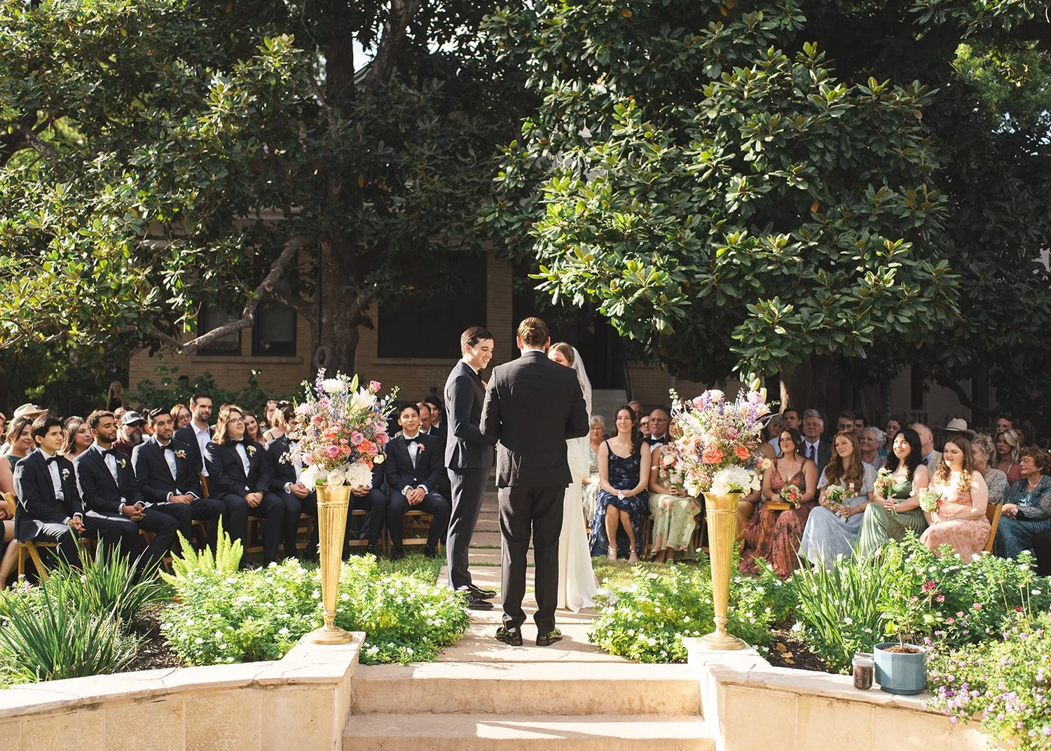 Wedding ceremony outdoors with bride and groom exchanging vows, surrounded by guests seated in a garden with large trees and floral arrangements in tall vases.