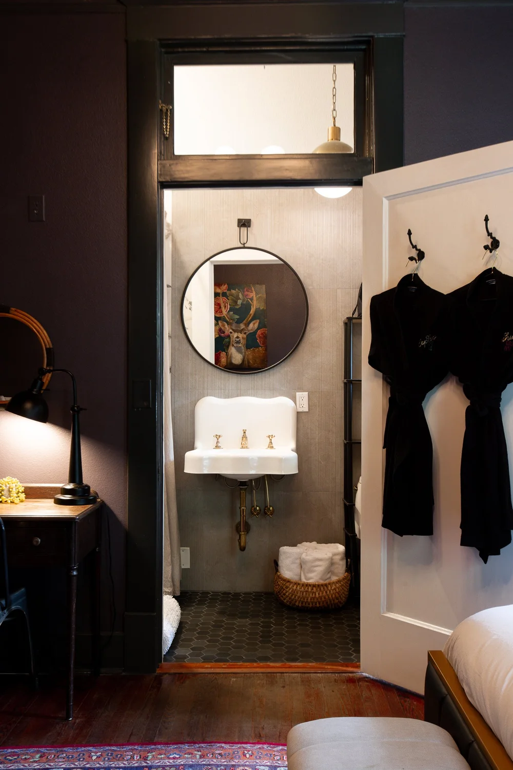 A small vintage bathroom with a white wall-mounted sink, a round mirror, a framed deer painting, and black hexagonal floor tiles. The bathroom is visible through a doorway from a bedroom with a bed, a nightstand, and a lamp.