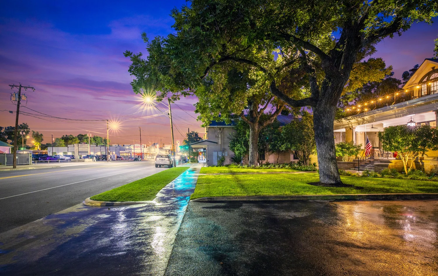 Nighttime street scene with wet sidewalk, lit streetlights, large trees, and a well-lit house with an American flag.