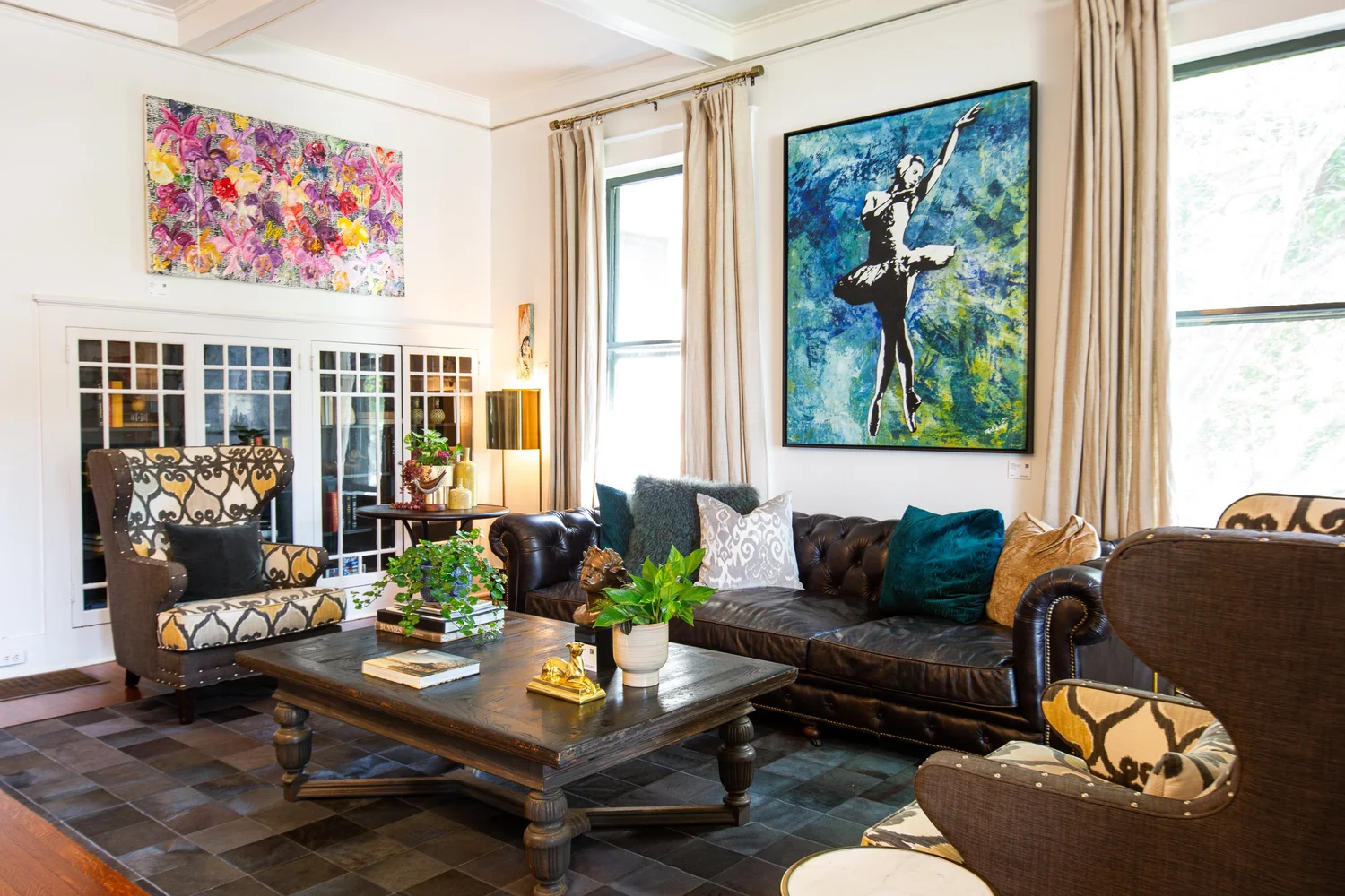 Living room with patterned armchairs, a tufted leather sofa with colorful pillows, a wooden coffee table with plants and books, two large windows with beige curtains, colorful artwork on white walls, a bookshelf, and a floor lamp.