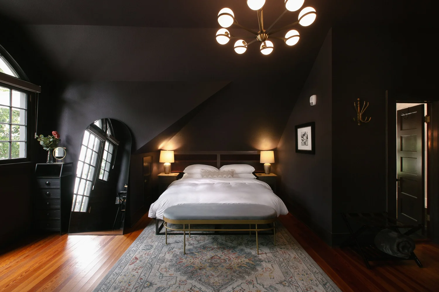 Dark-themed bedroom with black walls, white bedding, two bedside lamps, a gold bench at the foot of the bed, a large arched mirror, a window with black trim, and a modern chandelier.